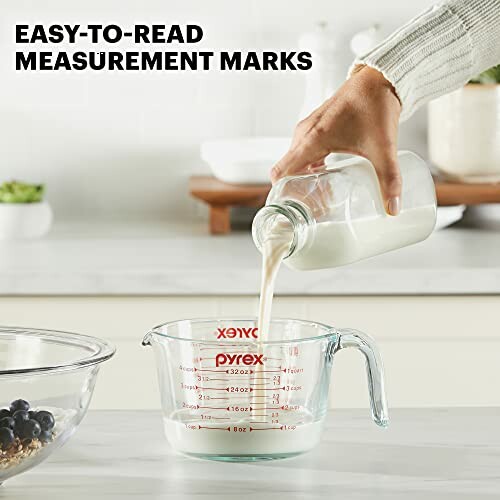 Hand pouring milk from bottle into a Pyrex measuring cup on a kitchen counter.